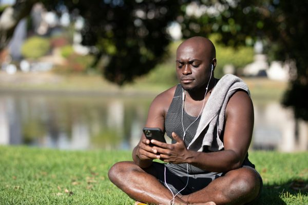 Bald man sitting in park using smartphone with earphones on a sunny day, showcasing a moment of leisure.
